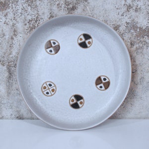 May include: A white ceramic plate with a geometric pattern of brown, black, and white circles. The plate has a textured surface.