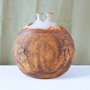 May include: A brown ceramic vase with two openings at the top. The vase has a white rim and a textured surface with a brown glaze. The vase has a round shape and is decorated with carved designs.