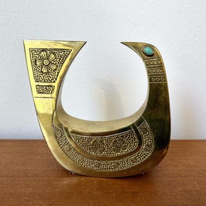 May include: A gold-colored metal bird sculpture with intricate floral designs and a turquoise blue gemstone accent. The bird is in a standing position with its head turned to the side.