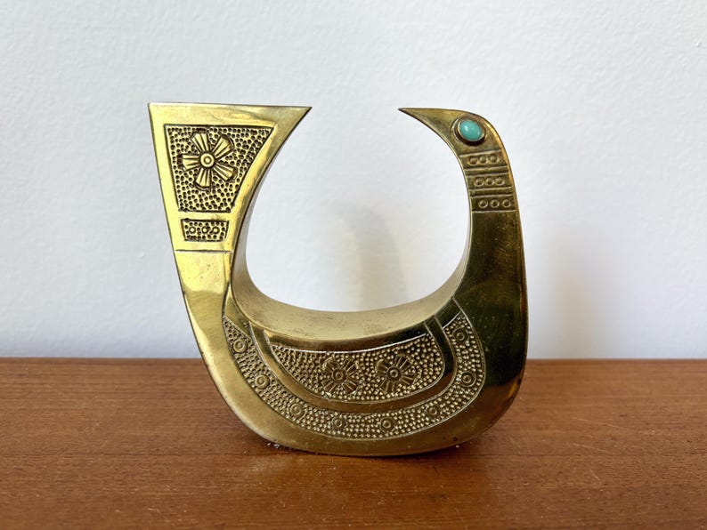May include: A gold-colored metal bird sculpture with intricate floral designs and a turquoise blue gemstone accent. The bird is in a standing position with its head turned to the side.