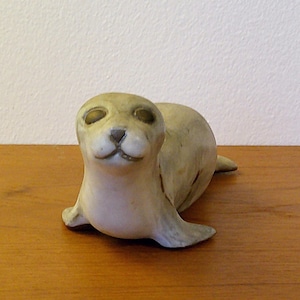 Puede incluir: Figura de foca de cerámica en color beige claro. La foca está representada en posición de descanso, con las aletas extendidas. La figura tiene un acabado liso y está colocada sobre una superficie de madera.