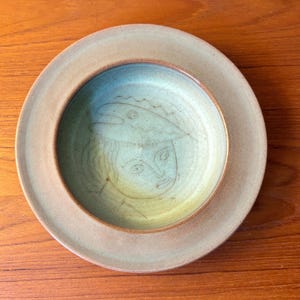 Puede incluir: Un juego de plato y cuenco de cerámica hechos a mano. El plato es de color beige claro, y el cuenco es de color azul verdoso claro con un diseño de cara esbozada. El conjunto está sobre una superficie de madera.