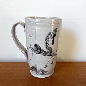 May include: A tall, ceramic mug with a seahorse design. The mug is light gray with a black and white seahorse illustration. The handle is curved and the mug sits on a wooden surface.