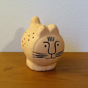 May include: A tan ceramic cat figurine with black facial features and a textured body. The cat has a whimsical design with a round body, upright ears, and a tail. The figurine is sitting on a wooden surface.