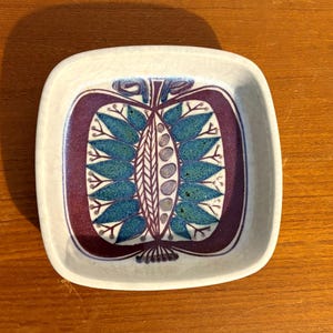 Puede incluir: Un plato cuadrado de cerámica blanca con un diseño estilizado de una fruta morada y verde sobre fondo blanco.