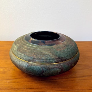 May include: A round, decorative bowl with a dark interior and a textured exterior. The bowl has a green, brown, and gold color scheme with a gold band around the middle. The bowl is sitting on a wooden surface.