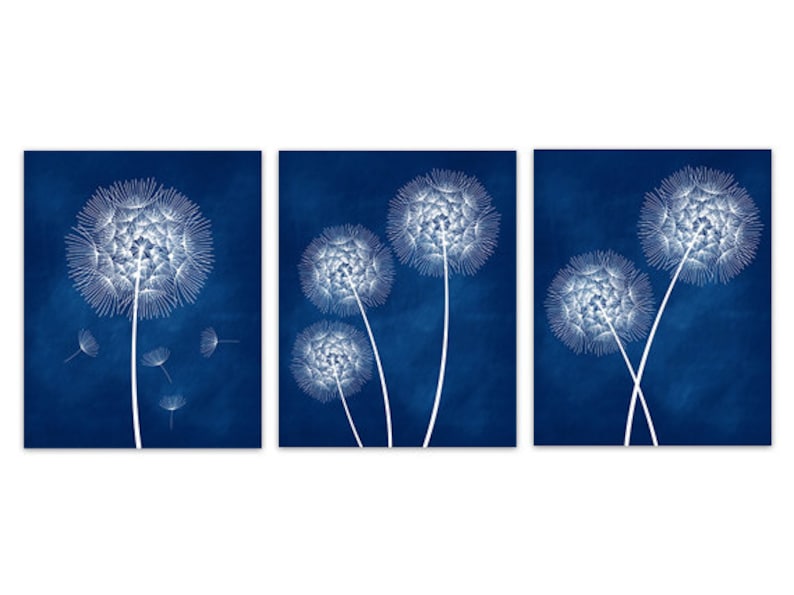 May include: Set of three wall art prints featuring white dandelion seed heads against a deep blue background. The dandelion illustrations have intricate details, with some seeds floating away. The artwork is a modern, nature-inspired design.