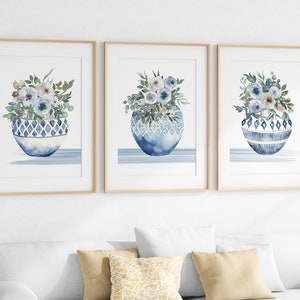 May include: Set of three framed watercolour prints featuring blue and white floral arrangements in blue and white patterned vases. The prints have a light blue background and are framed in natural wood.