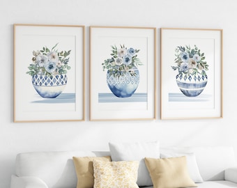 Puede incluir: Conjunto de tres impresiones de acuarela enmarcadas que presentan arreglos florales azules y blancos en jarrones con patrones azules y blancos. Las impresiones tienen un fondo azul claro y están enmarcadas en madera natural.