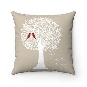 May include: A beige throw pillow featuring a white silhouette of a tree with two red birds perched on a branch. The tree has a white trunk and branches with white leaves.