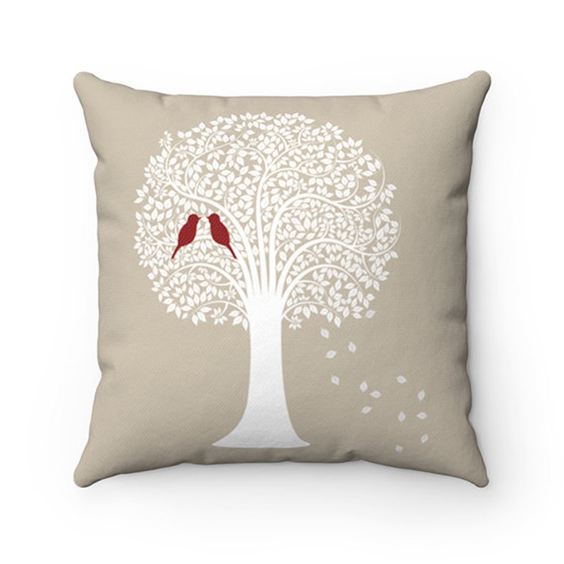 May include: A beige throw pillow featuring a white silhouette of a tree with two red birds perched on a branch. The tree has a white trunk and branches with white leaves.