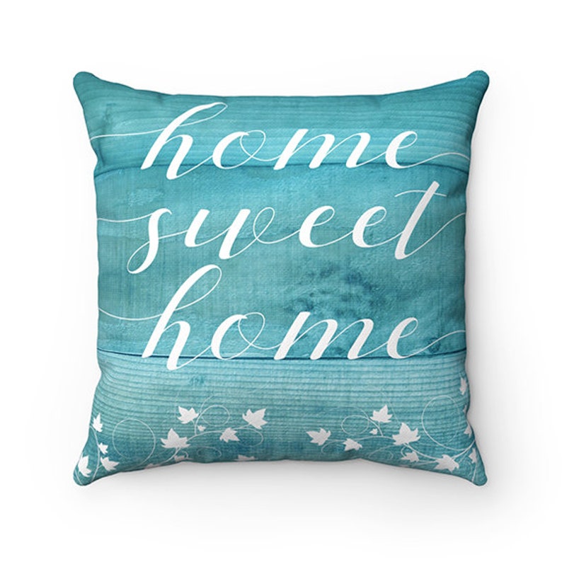 May include: A turquoise blue throw pillow with a white design that says "home sweet home" on a woodgrain background. The pillow has a white vine pattern around the edges.