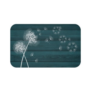 May include: Rectangular teal-colored decorative mat featuring a white dandelion design. Two large dandelions are on the left, with several seeds blowing away. The background has a wood grain texture.