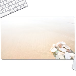 May include: A rectangular mousepad featuring a beach scene with sand and seashells. A white computer keyboard and mouse are in the frame. The mousepad has a light beige background with a cluster of seashells in the lower right corner.
