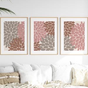 May include: Three framed floral art prints. Each print features a stylized flower design in shades of pink, brown, and grey against a cream background. The frames are a light wood colour, and the artwork is displayed above a white sofa.