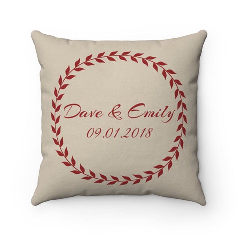 May include: A beige throw pillow with a red wreath design. The wreath has leaves and the text "Dave & Emily 09.01.2018" in the centre.