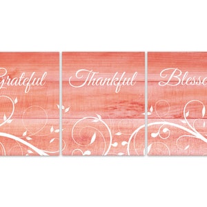 May include: Three wooden signs with a pink woodgrain background and white floral designs. The signs read "Grateful", "Thankful", and "Blessed".