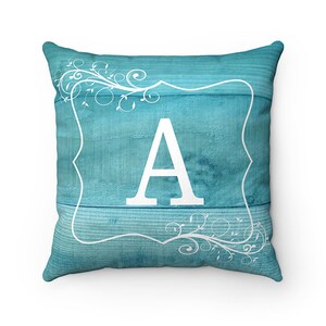 May include: A turquoise blue throw pillow with a white letter A in a decorative frame. The frame is surrounded by a white floral design.