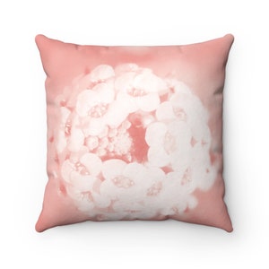May include: Square throw pillow with a floral design. The pillow features a soft, muted coral background with a close-up of white flowers. The flowers are clustered together, creating a textured appearance. The pillow is a square shape.