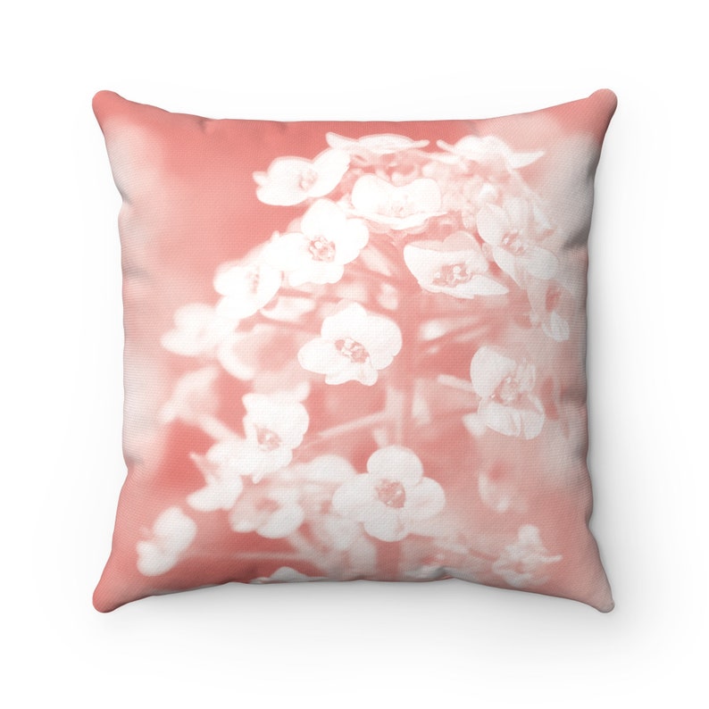 May include: Square throw pillow with a floral design. The pillow features a soft coral background with a close-up of white flowers. The flowers are clustered together, creating a delicate and airy aesthetic. The pillow is a decorative home accent.