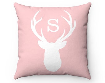 May include: Pink throw pillow with a white silhouette of a deer head with antlers and the letter "S" in the center.