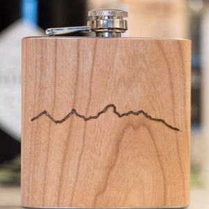 Pode incluir: Uma garrafa de madeira com uma tampa de prata e um design de cordilheira de montanhas gravado a laser.