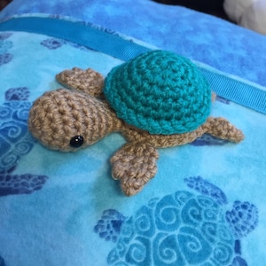 May include: A crocheted sea turtle toy with a brown body and a teal shell. The turtle is sitting on a blue and teal fabric with a printed pattern of sea turtles.