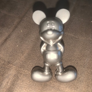 Puede incluir: Una figura plateada de Mickey Mouse. La figura tiene orejas grandes y redondas, una cara sonriente y pies redondeados. La figura está de pie con los brazos a los lados. El diseño general es liso y metálico.
