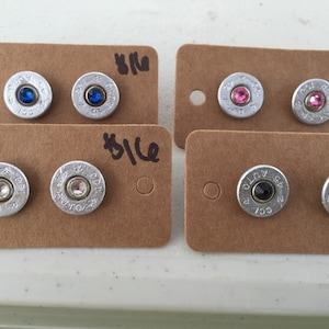 May include: Four pairs of stud earrings displayed on brown cards. The earrings are made from silver-colored bullet casings, each featuring a different colored gemstone: blue, pink, clear, and black. The cards have a small hole and handwritten text.