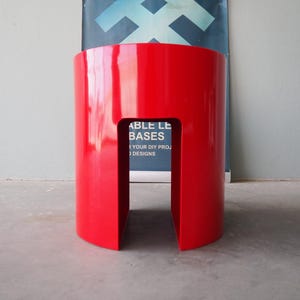 May include: A vibrant red, cylindrical stool with a rectangular cutout. The glossy finish reflects light, highlighting its smooth surface. The stool's design is modern and minimalist, suitable for various interior settings.