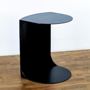 Peut inclure: Une table d'appoint noir mat dotée d'une structure en forme de C continu. L'objet se compose d'un plateau circulaire plat relié à une base circulaire identique par un panneau vertical incurvé, formant une silhouette unique, fluide et ouverte.
