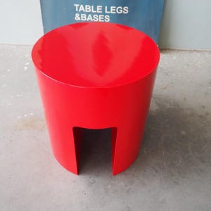 May include: A bright red, cylindrical stool with a cutout on one side. The stool has a glossy finish and a flat, circular top. The background includes a blue sign with the text "TABLE LEGS & BASES".
