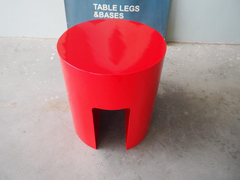May include: A bright red, cylindrical stool with a cutout on one side. The stool has a glossy finish and a flat, circular top. The background includes a blue sign with the text "TABLE LEGS & BASES".