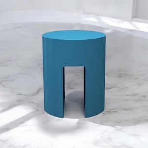 May include: A teal blue cylindrical stool with a rectangular cutout. The stool has a flat top and is made of a solid material. The stool is approximately 18 inches tall and 12 inches in diameter.