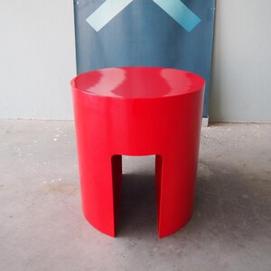 May include: A vibrant red, cylindrical stool with a cutout base. The stool has a glossy finish and a rounded top. The design is modern and minimalist. The stool is approximately 16 inches tall.
