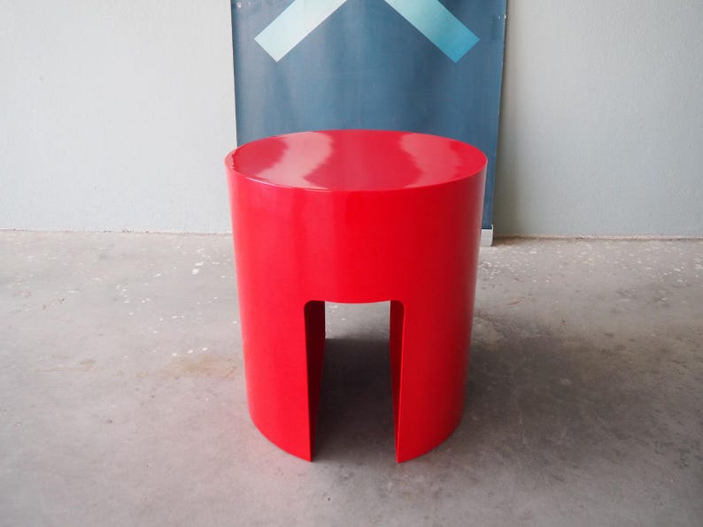 May include: A vibrant red, cylindrical stool with a cutout base. The stool has a glossy finish and a rounded top. The design is modern and minimalist. The stool is approximately 16 inches tall.