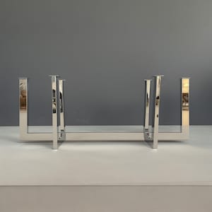Op de afbeelding: Een chromen metalen haardrooster met een rechthoekige vorm en een horizontale balk die de verticale balken verbindt. Het rooster is ontworpen om een ​​haard te beschermen tegen vonken en gloeiende kolen.