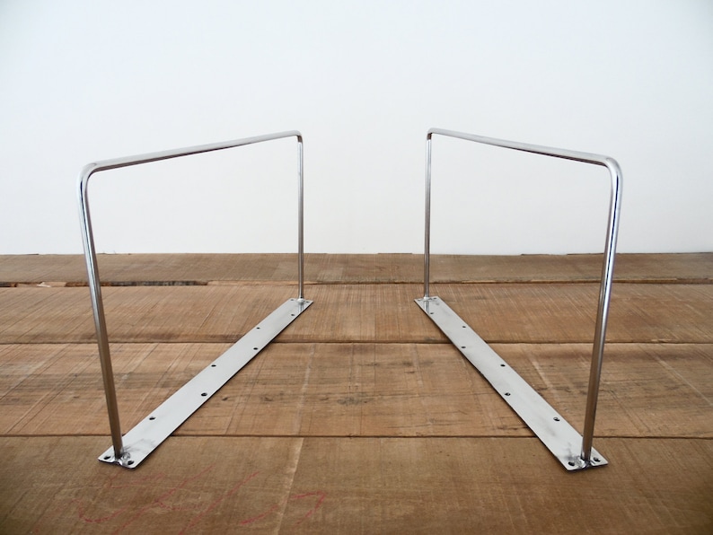 May include: Two silver metal shelf brackets with a flat base and a curved top. The brackets are placed on a wooden surface.