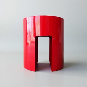 May include: A glossy, bright red cylindrical stool with a rectangular cutout. The stool has a smooth, reflective surface and a modern design. The cutout creates a handle-like opening. The stool is a vibrant accent piece.