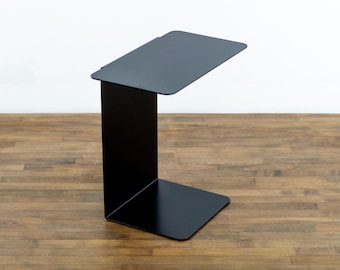 May include: A matte black C-shaped side table constructed from a single continuous piece of bent metal. The table features a flat rectangular top surface, a vertical support panel, and a flat rectangular base with rounded corners.