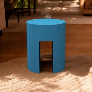 May include: A bright blue cylindrical side table with a rectangular cutout. The table is made of a solid material and sits on a beige rug. The table's design allows for storage within the cutout. The table is approximately 18 inches tall.