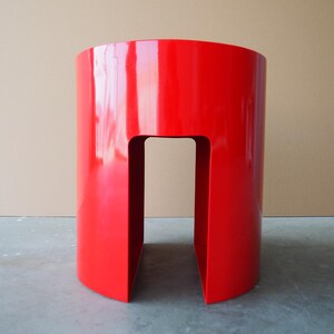 May include: A vibrant red, cylindrical side table with a glossy finish. The table has a cutout section in the center, creating an open space. The design is modern and minimalist, suitable for various interior settings.