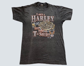 May include: Dark gray t-shirt with the text "I OWN A HARLEY NOT JUST A T-SHIRT" and a Harley-Davidson logo. The graphic is in shades of brown, orange, and white. The shirt has short sleeves and a crew neck.