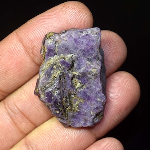 Peut inclure: Une pierre précieuse brute, naturelle, de couleur violette, avec une structure cristalline. La pierre présente un mélange de violets profonds et de lavandes plus claires, avec des touches d'or et de brun. La surface est texturée et irrégulière.