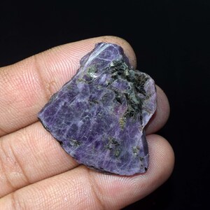 Può includere: Un pezzo grezzo a forma di cuore di pietra preziosa di charoite viola. La pietra ha un aspetto marmorizzato con inclusioni verde scuro e nere. La charoite è tenuta in una mano su uno sfondo nero.