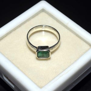 May include: A silver ring featuring a rectangular, emerald-cut green gemstone set in a smooth metal bezel. The band is polished silver and includes a small 925 hallmark stamped on the inner surface, indicating sterling silver composition.