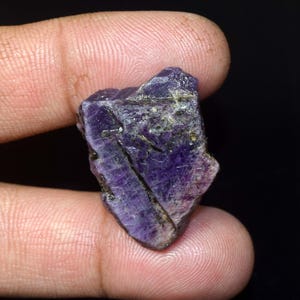 Puede incluir: Una gema de amatista natural en bruto, que muestra un tono púrpura intenso con vetas e inclusiones más claras. El cristal en bruto se sostiene entre los dedos sobre un fondo oscuro, mostrando su forma y textura irregulares.