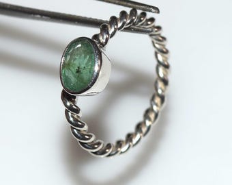 May include: A silver ring with a twisted band design, featuring an oval-cut emerald gemstone. The emerald has a vibrant green color and is set in a bezel setting. The ring is held by a pair of tweezers.