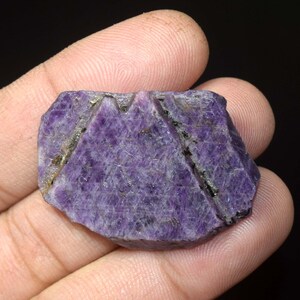 Op de afbeelding: Een ruw geslepen, natuurlijke paarse edelsteen met een kristallijne structuur. De steen heeft een diepe violette tint met lichtere strepen en donkere insluitsels. De randen zijn ongelijkmatig en het oppervlak heeft een textuur.