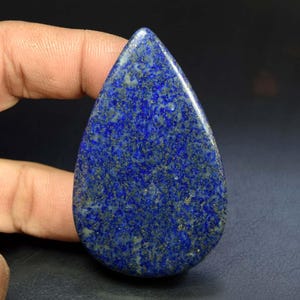 May include: A teardrop-shaped lapis lazuli gemstone cabochon featuring a deep blue base color with scattered golden pyrite inclusions and white mineral flecks throughout the polished, smooth surface of the stone.